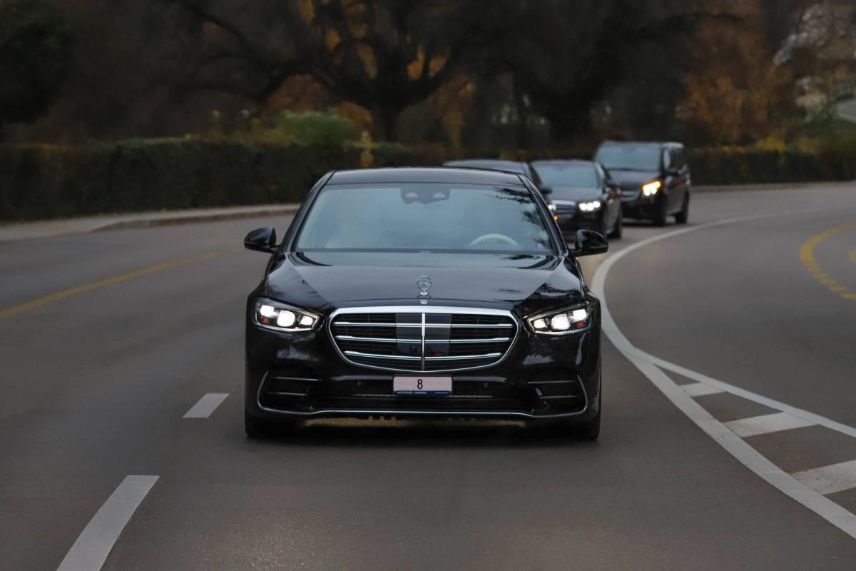 A row of five black luxury cars parked side by side on a paved road, each with an individual in formal attire standing beside it, set against a backdrop of greenery, trees, and a partially visible building.