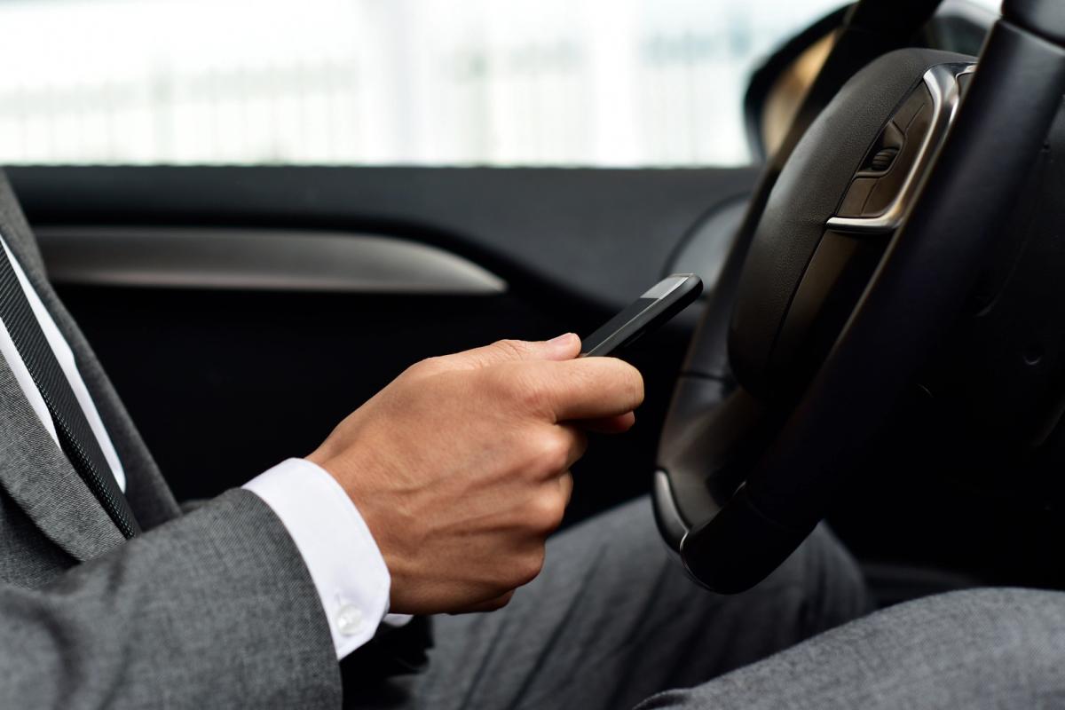 Professional chauffeur in a tailored suit using a smartphone in the driver’s seat of a luxury rental car in Switzerland, highlighting the car’s elegant interior and modern features.