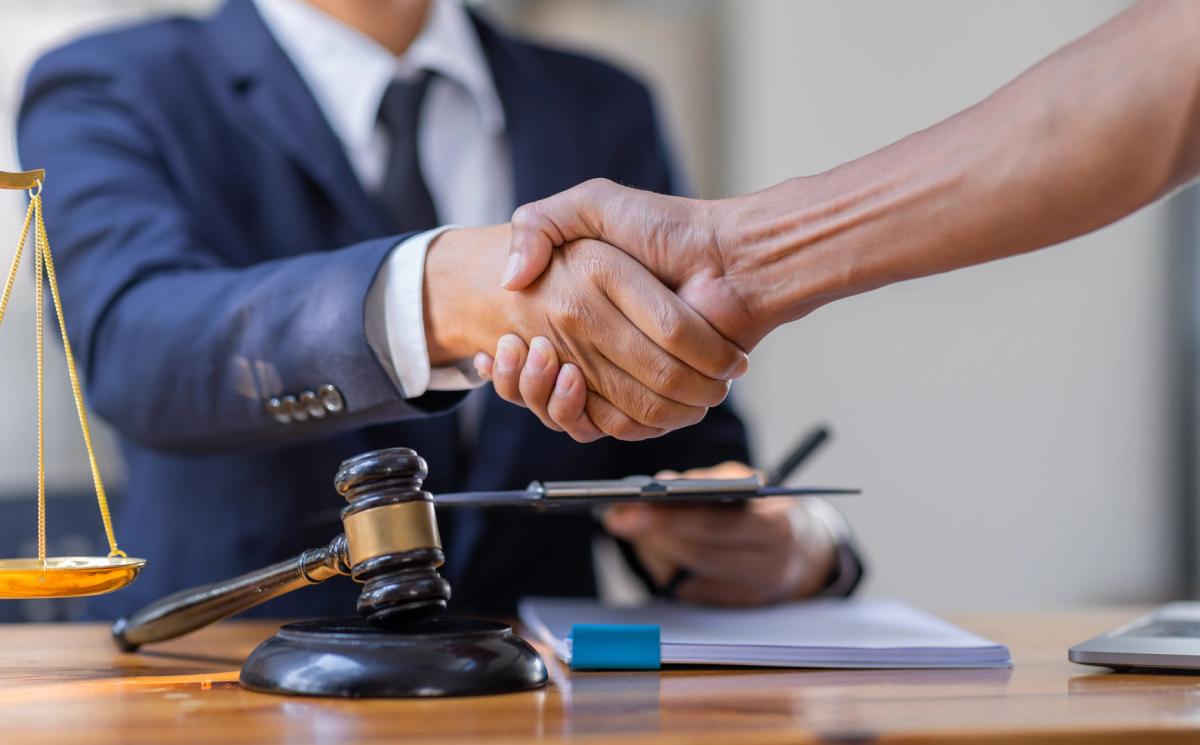 Two professionals shaking hands across a wooden table in a well-lit office, with a gavel, sound block, and golden scale of justice visible, indicating a legal context or agreement.