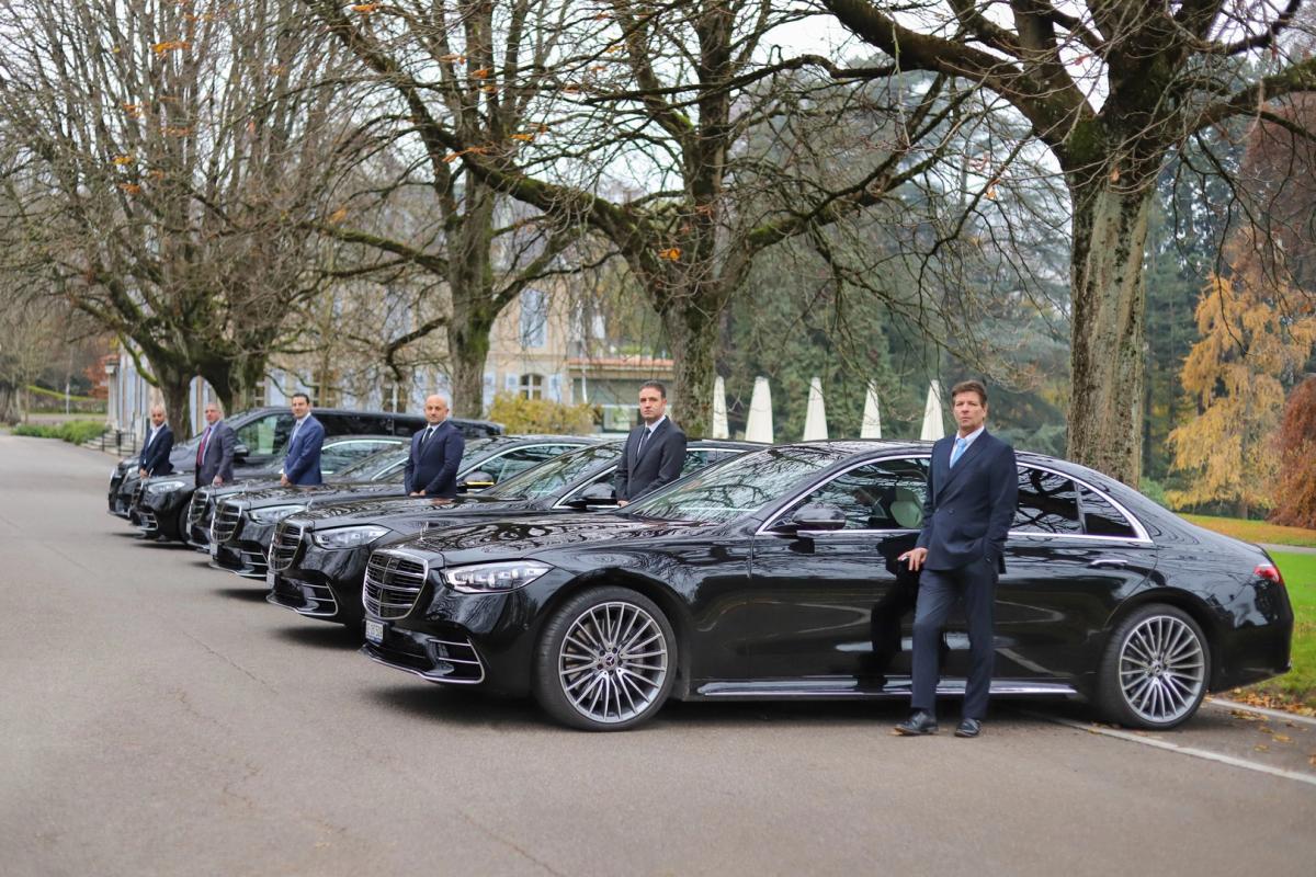 A row of five black luxury cars parked side by side on a paved road, each with an individual in formal attire standing beside it, set against a backdrop of greenery, trees, and a partially visible building.