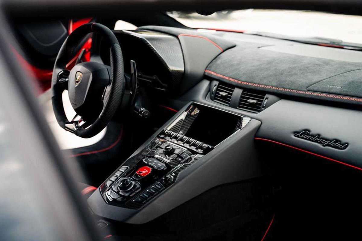 Interior view of a luxury Lamborghini showcasing the sleek black steering wheel with the iconic logo, red and black leather seats, and a high-tech center console. The dashboard is inscribed with 'Lamborghini', emphasizing the brand's prestige. The red stitching details enhance the aesthetic appeal, making it an ideal choice for luxury car rentals in Switzerland.