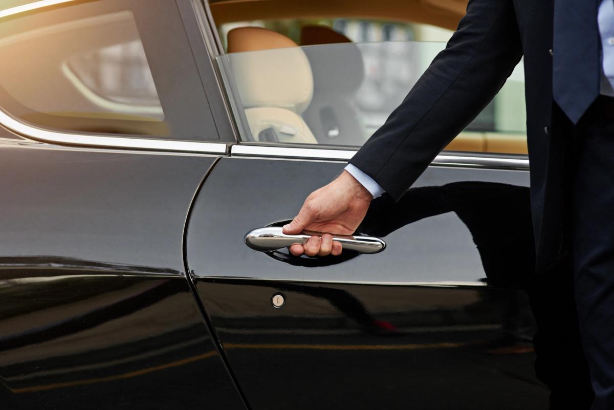 A person in a dark suit opening the door of a sleek, black car. The car's luxurious and spacious interior with light-colored seats is partially visible through the window. The image is set in an outdoor setting during daylight hours, perfect for showcasing the elegance of the car available for luxury rental in Switzerland.