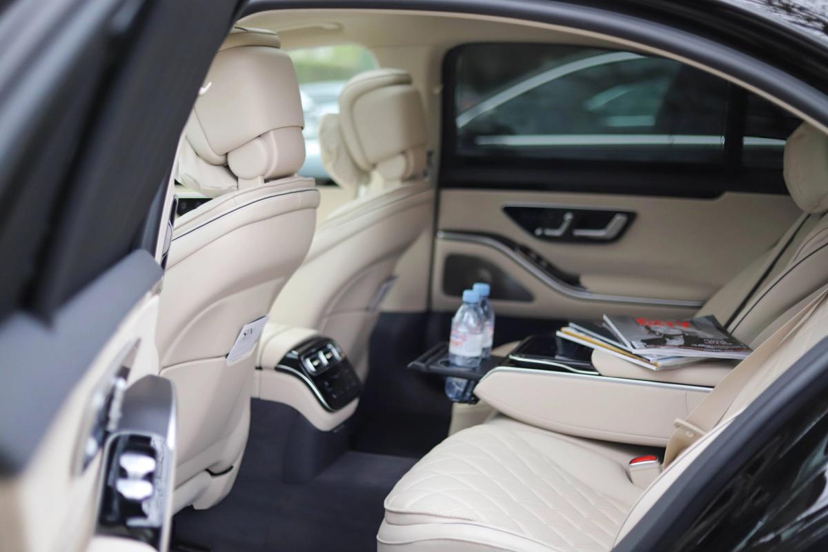 Interior of a car showcasing cream-colored, luxurious leather seats with detailed stitching. A bottle of water and magazines are placed on one of the seats. The interior design includes sleek black and cream contrasts, with modern controls visible on the rear armrest. Ideal for luxury car rental services.