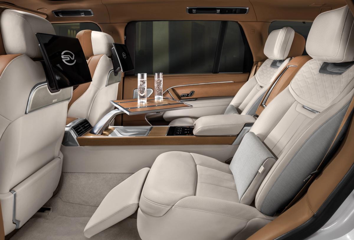 Luxurious and spacious car interior featuring beige leather seats with intricate stitching patterns. The polished wooden center console includes cup holders, controls, and small table that extends out towards the rear seats. Two champagne glasses are placed on the table, indicating a level of opulence.